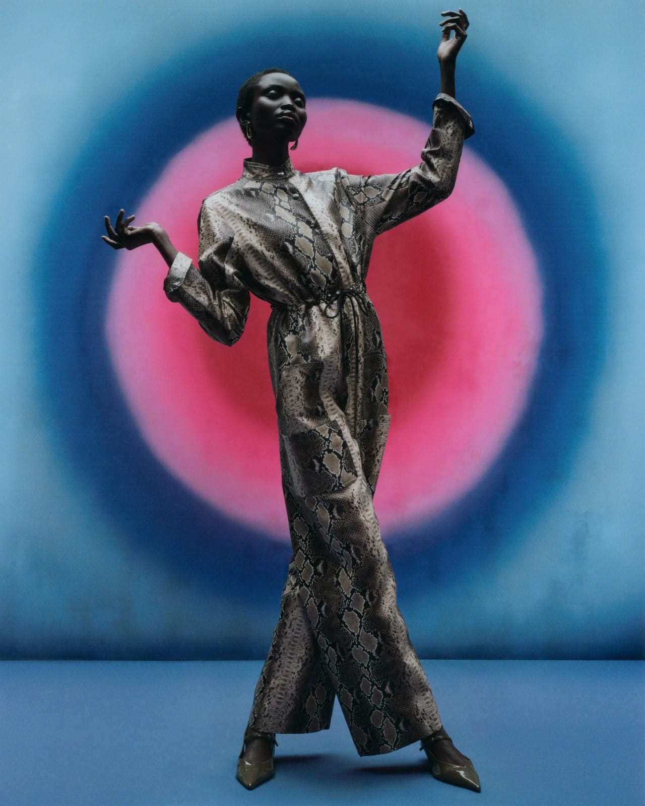 Fashion editorial: a model in a snakeskin two-piece with arms raised against a hand-painted blue and pink concentric circle backdrop