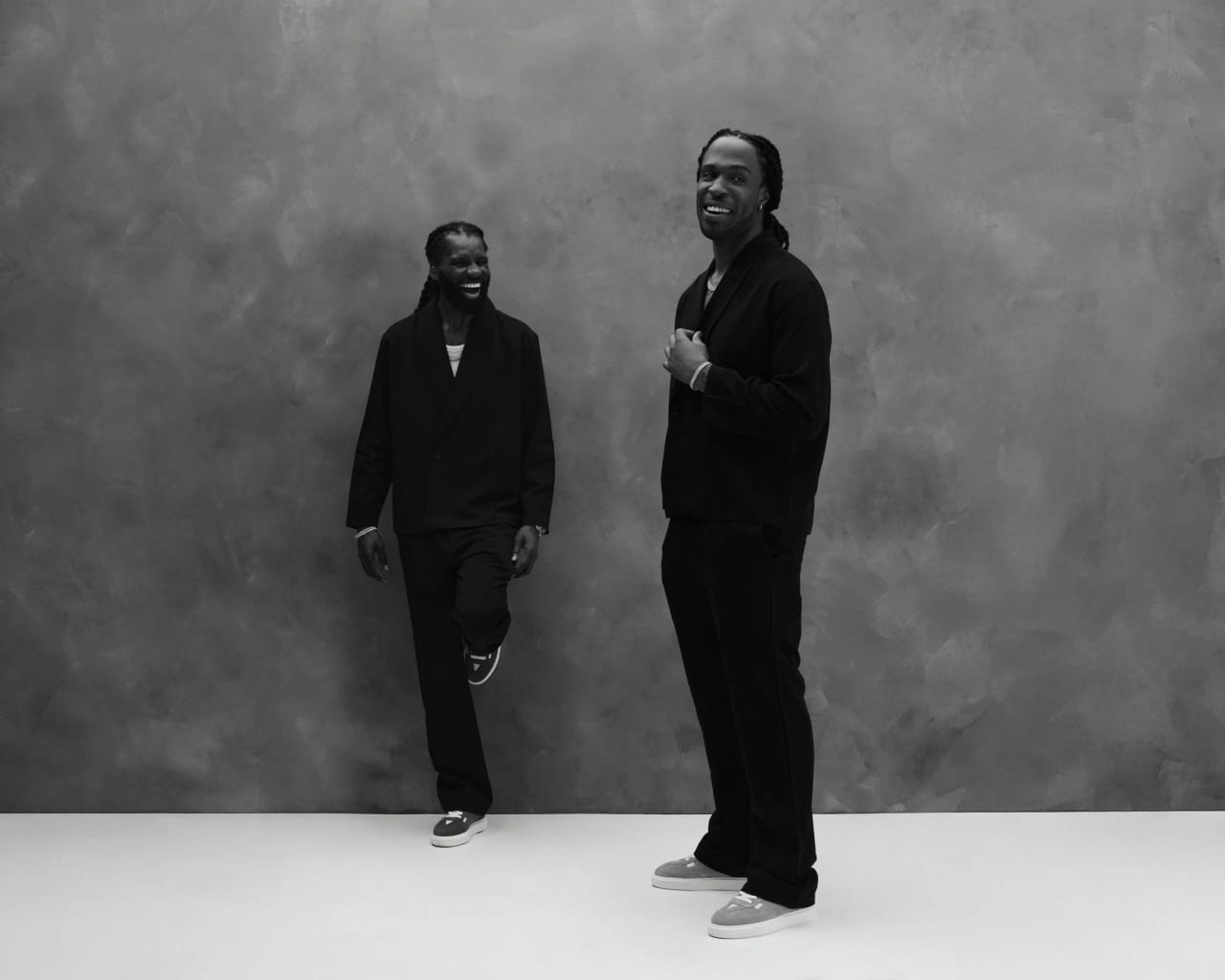 Two artists photographed together in black on a mottled grey backdrop, mid-laugh